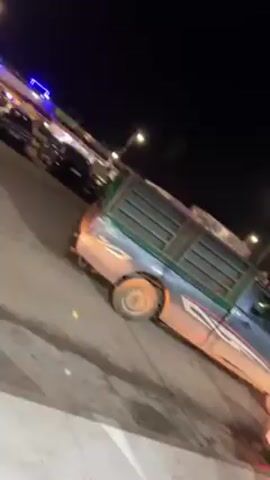 The video shows a street scene at night, likely in a parking area near a food vendor. A silver pickup truck with a green cage-like structure in its bed is parked on the left side of the frame. There are other cars parked nearby, including a red truck in the background. The camera is positioned under the awning of a food stand, which has "Cremio" branding visible. The lighting is provided by street lamps, and there appears to be some smoke or fog in the air. There are people in the area, but they are not clearly visible.