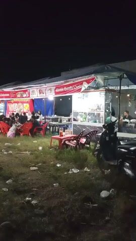 The video shows an outdoor night scene of a food stall area and a carnival ride. In the foreground, there is a grassy area littered with plastic bottles and debris. A dark-colored motorcycle is parked on the right. To the left, a food stall with a red and white striped awning is visible. Several red plastic chairs are arranged around tables, with people seated and eating. A sign above the stall reads "Catir Banda Unik". Further to the left, another stall with a banner displaying food images is present.

In the mid-ground, a brightly lit Ferris wheel, a carnival ride, dominates the scene on the left. Next to it, a food or game stall is set up under a tent. Several people, including women in hijabs and children, are walking around this area.

The lighting is dim, with artificial lights from the stalls and the Ferris wheel illuminating the scene. The overall atmosphere appears to be that of a local night market or fair.