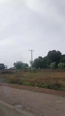The video shows a wide-angle view of a semi-rural or rural landscape in Pasere, Nigeria, on an overcast day. The camera moves forward, suggesting a vehicle is in motion.

In the foreground, a road with a broken, somewhat paved surface and noticeable potholes is visible. One large puddle of water is visible in a pothole. As the camera moves, the road appears to transition to a dirt or unpaved track on the right side.

The mid-ground consists of patches of green grass and dirt, with several scattered trees. To the left, a few simple, distant buildings or houses are partially visible amongst the trees. On the right, a tall utility pole with power lines extends across the frame. Briefly, around the 0:03-0:04 mark, another pole-like structure, possibly a sign or a stand with a solar panel on top, appears near the road.

The background is dominated by a light, overcast sky, with more trees and distant structures barely visible on the horizon. There are no people visible, and no discernible text is present in the video.