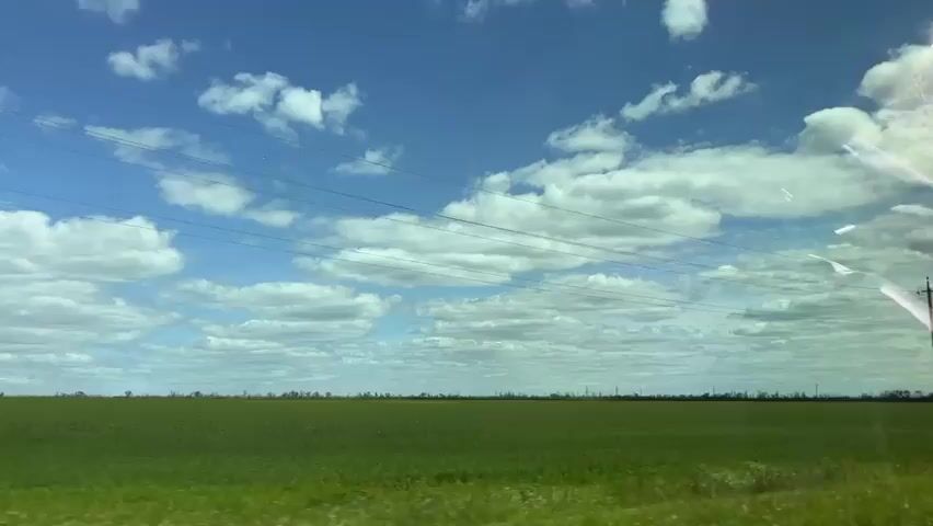 The video depicts a landscape viewed through a car window. The sky is a bright blue with scattered white clouds. The horizon features a flat green field stretching as far as the eye can see. Power lines run horizontally across the frame, suspended by utility poles that punctuate the landscape. A road sign is partially visible. The video seems to capture a scene of rural travel on a clear day.