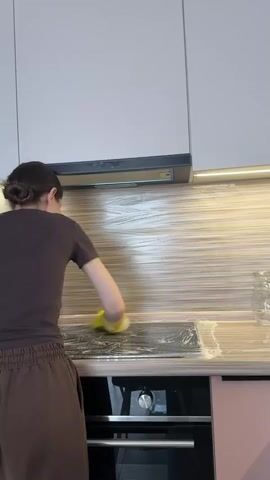 A person wearing a brown t-shirt and brown sweatpants with their hair pulled back in a bun cleans a kitchen counter with a yellow sponge. The person is standing in front of a black stovetop that has a white foamy substance on it. The kitchen has wooden cabinets and a grey backsplash. The person is wearing yellow gloves.
