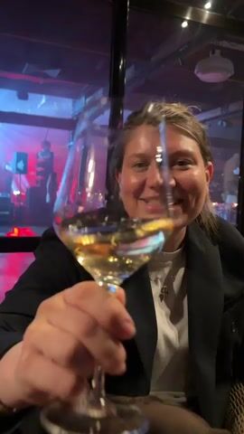 A woman smiles and holds up a glass of white wine. The scene is dimly lit with purple and red lights in the background suggesting a bar or event space. In the background is a stage with figures, possibly musicians. The woman is wearing a black blazer over a white shirt and a silver necklace. The focus is on her and the glass of wine, blurring the background.