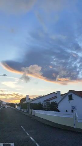 A vertical shot shows a street lined with white houses and a few trees. The sky above is a blend of blue and white clouds, with a prominent lenticular cloud formation illuminated by the setting sun. The road is paved and has white lines. No people are visible in the video. The overall scene depicts a quiet, residential street at dusk.