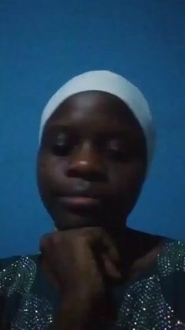 The video features a close-up shot of a young Black woman from the chest up, positioned against a plain blue wall. She is wearing a white head covering that fully covers her hair and a dark-colored top or dress adorned with intricate patterns of shiny, possibly sequined or rhinestone, embellishments across the shoulders and chest.

Her face is well-lit, showing a natural, somewhat dewy complexion. Throughout the video, she exhibits a range of subtle expressions and gestures. At the beginning, she looks contemplatively to the side with her hand near her chin, then shifts her gaze towards the camera while making a slight pout with her lips. She brings her hand to her chin multiple times, often with a thoughtful or slightly serious expression, occasionally looking down or to the side.

At various points, she makes small mouth movements, including clicking sounds. Around the 0:19 mark, she makes an "okay" hand gesture (thumb and forefinger touching, other fingers extended). Later, she shows five fingers with her hand and makes snapping motions. Towards the end of the clip, she briefly points to the side of her right eye with her index finger, then smiles faintly. Her expressions fluctuate between serious, pensive, and briefly smiling, as she looks directly into the camera or slightly away. The background remains constant throughout the video.