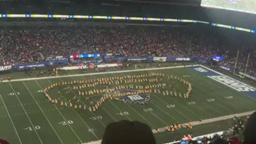 The video shows a marching band forming the shape of a guitar in the middle of a football field. The band is wearing yellow and black uniforms and is surrounded by a large crowd of spectators. The field is marked with the words "GIANTS" at each end. The stadium is full and appears to be a large, professional venue.