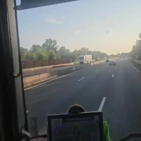 The video shows a highway with several vehicles, including a large white truck, a black SUV, and several red fire trucks with flashing blue lights. The fire trucks appear to be moving in the same direction as the other vehicles, suggesting a convoy or a response to an incident. The environment is sunny with trees lining the side of the highway. The video is shot from the perspective of a vehicle traveling on the highway.