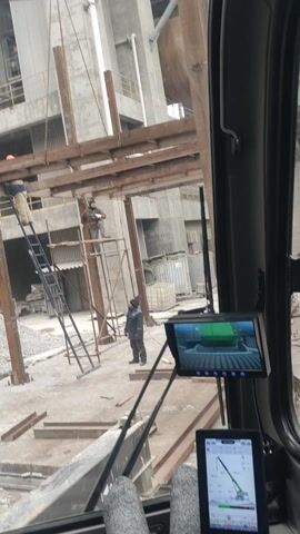 The video shows a view from inside the cab of a crane, looking out at a construction site. Several construction workers are visible on scaffolding, some of whom are using a ladder to access higher areas. The crane operator's legs, clad in jeans and light-colored socks, are visible. Two screens are mounted on the dashboard, displaying information, including a visual of the crane's arm and what appears to be its weight distribution.  The crane operator appears to be at a standstill.