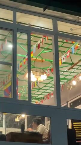 The video shows the outside of a building at night. Through large glass windows on the upper level, one can see a person sitting at a table inside a room, which is decorated with numerous international flags hanging from the ceiling. The interior is lit by overhead lights. The lower level of the building is marked with signs, including one that reads "VILLAGE BOUTIQUE" and "FREE TIBET". The camera pans to the left, revealing more of the building and storefronts along a street.