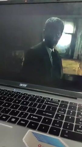 A laptop sits on a surface, playing a movie scene. A Black man in a dark suit and light shirt stands indoors, near a window. The scene changes to an open refrigerator with food items inside. The laptop is an Acer brand. A stack of cards is visible on the laptop's surface.