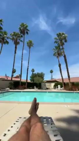 The video shows a first-person perspective of a person relaxing on a lounge chair next to a swimming pool on a bright, sunny day. The person's bare legs are visible, crossed at the ankles, extending towards the pool. The clear blue swimming pool occupies the mid-ground of the scene.

Tall palm trees with green fronds are prominently featured, standing against a clear blue sky with a few wispy white clouds. Several buildings with light-colored walls and red-tiled roofs are visible behind the pool and palm trees, suggesting a resort, hotel, or residential community. The overall atmosphere appears peaceful and relaxing, indicative of a vacation or leisure activity in Palm Desert, United States. There is no visible text in the video.