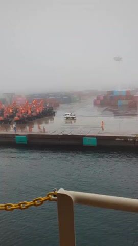 The video shows a foggy harbor scene. In the foreground, a yellow chain is visible across the bottom of the frame. Beyond the chain, dark blue water is seen. In the middle ground, a dock or pier extends into the water. On the dock, a line of orange excavators is parked. Behind the excavators, stacks of shipping containers in various colors are visible, partially obscured by fog. A white vehicle with a light colored canopy is driving on the dock between the excavators and the containers. One person in a blue shirt is standing near the excavators, and another person in an orange vest is standing further down the dock. The overall atmosphere is overcast and hazy due to the fog.