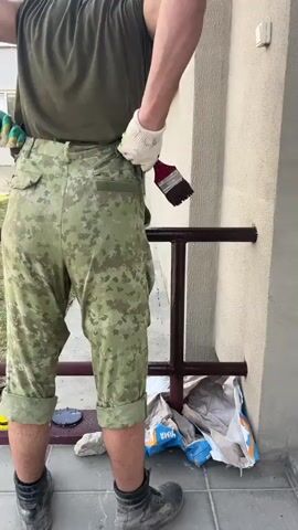 A person is shown from the back, wearing a dark green t-shirt, camouflage capri pants, and black boots. They are also wearing white work gloves. The person is standing on a tiled surface next to a light-colored building. A dark metal railing is attached to the building. The person is holding a brush and appears to be painting or applying something to the railing. Several bags of building material are on the ground near the railing, and a yellow bucket is also visible. In the background, there is a multi-story building with windows and an air conditioning unit. The overall scene suggests an outdoor maintenance or repair task. The time of day appears to be daytime, and the weather seems to be fair. There is no discernible text in the visible portion of the media.
