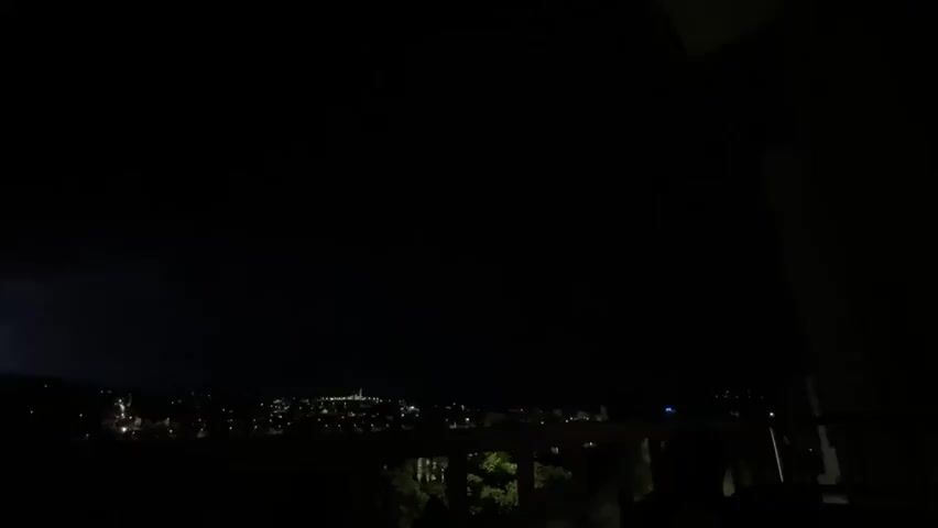 A nighttime scene is visible, apparently taken from a balcony.  The view overlooks a dark city landscape with visible clusters of lights marking buildings and streets. A person’s voice can be heard reacting with shock or surprise, exclaiming "What the fuck." The foreground shows railing and possibly some foliage, all mostly silhouetted against the night. The sky is dark.