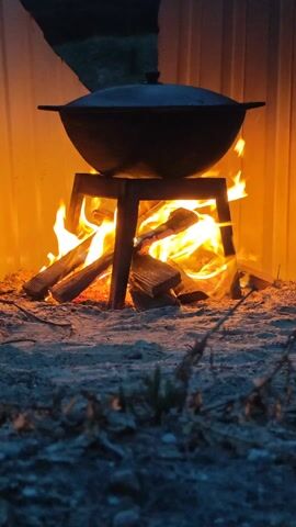 A close-up shot shows a large, dark, round pot resting on a short, four-legged metal stand. Beneath the pot and stand, a wood fire burns brightly, casting an orange glow on the scene. The fire is built on a bed of sand or dirt, with pieces of wood arranged to fuel the flames. Behind the fire and pot is a corrugated metal wall. A hand reaches into the frame and removes the lid of the pot, then replaces it. The overall lighting suggests it is either dusk or dawn, with the fire providing the primary source of illumination. The ground in the foreground is partially covered with dried grass.