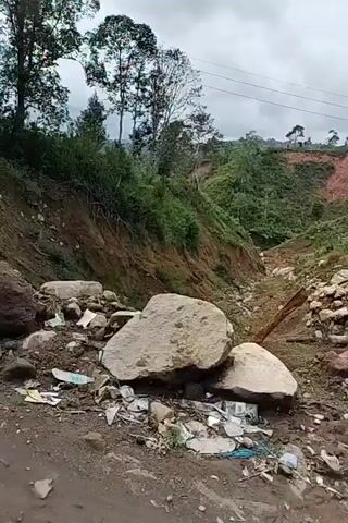The video shows a damaged landscape with debris and a landslide. The ground is muddy and uneven, with rocks and trash scattered around. A large boulder is visible in the foreground. The landslide has exposed the earth, creating a raw scar on the hillside. Trees and vegetation are growing on the slopes, but the damage from the landslide is evident. The sky is cloudy, suggesting overcast weather. The video appears to be taken from a moving vehicle, as evidenced by the slight motion blur and the perspective. The overall impression is one of a natural disaster and its aftermath.