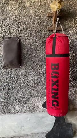 A red punching bag hangs from a chain attached to a wooden bracket mounted on a textured, gray wall. To the left of the punching bag, a small, dark rectangular object is also attached to the wall. The punching bag has black straps and the word "BOXING" printed vertically in large black letters, along with smaller text below. A dark, silhouette-like object rests at the base of the punching bag. The scene appears to be indoors, possibly a gym or training area.