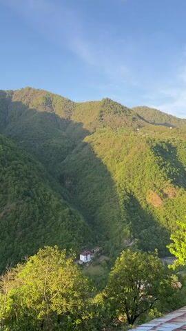 The video shows a view from a roof, looking out over a valley of green hills and mountains under a blue sky with wispy clouds. The hills are densely covered in trees. A small building can be seen nestled in the valley. The camera pans slightly to the right, revealing more of the landscape and the tiled roof from which the video is taken. In the background, some snow-capped mountains can be seen in the distance.  An indistinct voice can be heard in the background.