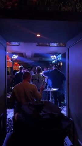 The video shows a band playing live music in a dimly lit establishment. The band members are positioned towards the back of the room, with the drummer in the foreground, facing away from the camera. The other band members, including guitarists and a singer, are facing the audience. The lighting shifts between vibrant purple and green hues, creating a dynamic atmosphere. The walls are adorned with patterned wallpaper, and ceiling lights provide additional illumination. In the background, patrons can be seen gathered, enjoying the performance. The overall scene suggests a lively evening of entertainment in a bar or club setting. The time of day is likely evening or night, given the artificial lighting and the nature of the event. There are no visible signs of weather or specific location cues beyond the general bar/club ambiance. The mood conveyed is energetic and engaging.