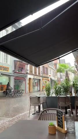 The video shows a street scene with several shops on the left side. A person on a bicycle is riding down the street, which is wet from the rain. In the foreground, there are outdoor tables and chairs, suggesting a cafe or restaurant setting. The overall atmosphere is rainy and overcast.