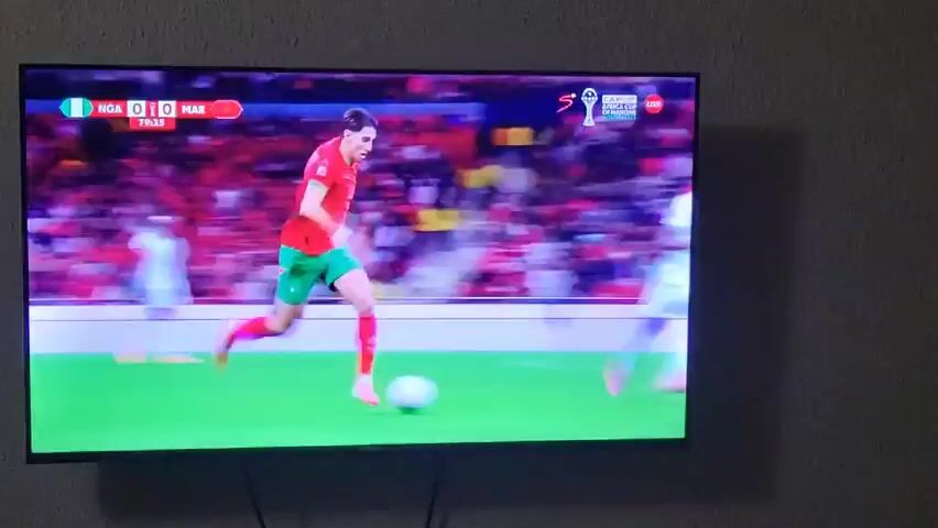 A television screen displays a soccer game, Nigeria versus Morocco. The score is tied 0-0. The time remaining in the game is 79:39.  Several players in red and green uniforms are pursuing the ball, while players in white uniforms attempt to defend. The game is being played in front of a packed stadium of spectators.  A shadow cast by the television is visible on the adjacent wall.