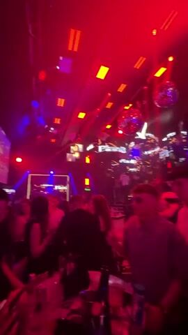 The video shows a crowded nightclub. The room is lit with red and blue lights and contains multiple disco balls. There are many people dancing and socializing. Some are standing at tables.  A large screen displays the words "PlanB".