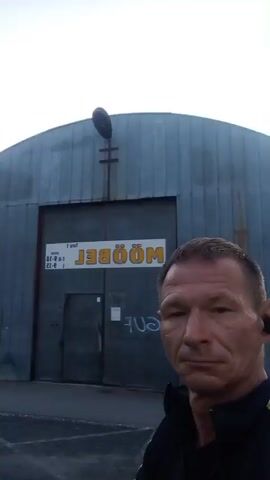 The video shows a man in the foreground, facing the camera, with a large, Quonset-hut-style building behind him. The building appears to be made of corrugated metal. There is a large set of double doors on the building, with a sign that is mostly obscured by the man's face. The sign has some text on it, but it is difficult to read. The man is wearing a black jacket with yellow and white accents. He has short, light-colored hair and is wearing black earbuds. The sun is shining brightly, casting lens flares across the video, and there is some vegetation visible to the right of the building. The ground in front of the building appears to be asphalt. The video is shaky, suggesting it may have been filmed with a handheld device.