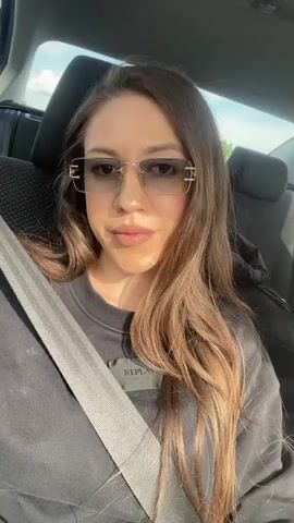 A woman in sunglasses and a black Replay sweatshirt is sitting in the passenger seat of a car, wearing a seatbelt. Her hair is long and brown with lighter highlights, and she has a full face of makeup. The car's interior is visible behind her. The video is shot from a slightly low angle, and the lighting is bright. Music is playing in the background. The woman turns her head slightly and smiles at the camera.