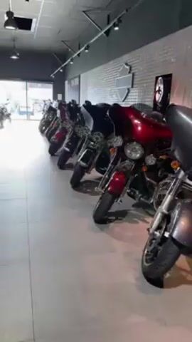 The video shows the interior of a Harley-Davidson motorcycle dealership. The camera pans across rows of motorcycles, displaying models in black, red, and other colors. The motorcycles are parked neatly in two rows. The building appears to be well-lit with a gray floor and a dark gray ceiling. A large window provides natural light in the front. On the walls, there are Harley-Davidson signs and decorations.