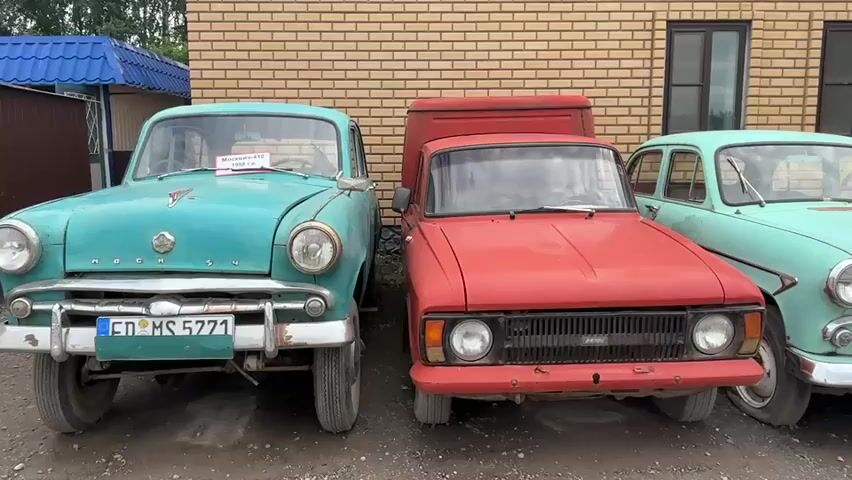 This media file shows a line of vintage cars parked outside a brick building. The cars are of various colors, including teal, red, yellow, blue, and pink. They appear to be Soviet-era models, with designs characteristic of the 1950s and 1960s. The vehicles are parked in a lot, with a gravelly surface in the foreground. Behind the cars, the brick building has several dark windows. A blue awning is visible on the left side of the frame.

The scene appears to be set during the daytime, under overcast skies. The cars are the primary subjects, and they are all stationary, suggesting they are part of an exhibition or a private collection. There are no people visible in the frame. The location is identified as Bol'shoi Mashlyak, Russia, which provides geographical context for the types of vehicles displayed.

Visible text includes license plates on some of the cars. For instance, the first teal car has a license plate with the text "ED MS 5771". Another teal car has a plate that reads "84-35 ТБГ". A yellow car's license plate is partially visible. There is also a small white sign on the windshield of the first teal car that appears to have Cyrillic text, possibly indicating the car's model and year. The overall impression is one of nostalgia and appreciation for automotive history.
