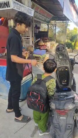 A young child wearing a backpack stands next to a motorcycle with its seat open. The motorcycle is parked on the side of a road in front of a small shop. The shop appears to be a general store or convenience store, with shelves stocked with various items visible through its open front. Further down the road, there are trees, houses, and other buildings. The sky is clear and blue, suggesting it is daytime. The overall scene depicts a common rural or semi-rural setting.
