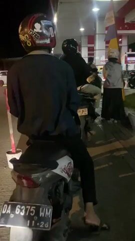 The video is taken at night at a gas station, identified by the "PERTAMINA" signage. In the foreground, a person on a scooter is partially visible from the back, wearing a dark jacket, black pants, and flip-flops. The scooter has a white and red sticker. Behind this person, another scooter with a rider wearing a black helmet is seen. Further back, at the gas pumps, there are several people and motorcycles. One person is wearing a light-colored jacket and a black skirt. A flag with colored stripes is visible near the gas pumps. The overall lighting suggests it is evening or nighttime, with artificial lights from the gas station illuminating the scene. A silver SUV is parked on the side of the road in the background of the second clip.