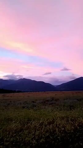 The video shows a landscape with a field in the foreground and mountains in the background. The sky is a vibrant pink and purple, indicative of a sunset or sunrise. The field appears to be covered in dry grass. The mountains are dark silhouettes against the colorful sky. There are clouds in the sky, some of which are tinged with pink and orange. The overall scene is serene and picturesque.