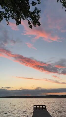 The video shows a sunset over a lake with a wooden dock in the foreground. The sky is a mix of blue and orange with thick pink and orange clouds. There are trees lining the opposite shore of the lake. A tree branch with leaves hangs in the upper left corner of the frame. A snippet of music plays in the background.