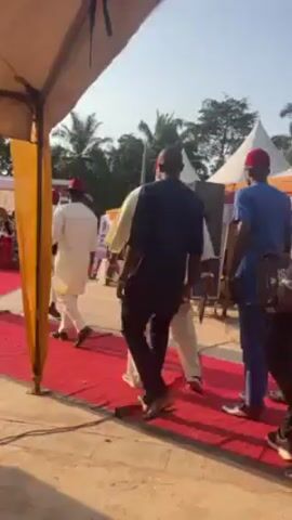 The video shows a gathering of people on a red carpet outdoors. A man in a black shirt and cap is speaking into a microphone. Several individuals wearing traditional attire are visible. The event appears to be taking place under a tent with a backdrop for photographs. There are trees and buildings in the background. Some people are walking and others are stationary, observing the speaker.