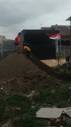 A group of people are on a pile of dirt, in front of a building. There is a flag hanging from the building. The sky is cloudy.