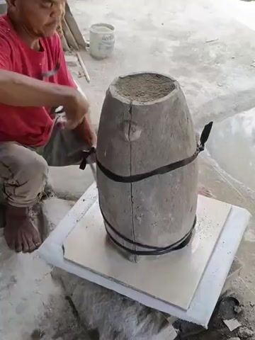 A middle-aged Asian man is shown in this medium shot, sitting barefoot in a squatting position on a concrete floor, next to a vertical, barrel-shaped mold. He is wearing a red t-shirt with a "Volcom" logo and camo-patterned pants. He is securing the mold, which appears to be made of cement or plaster, with black straps, tightening one of the straps with a hand tool. The mold is in two pieces, held together by the straps. It stands on a base made of a tile sitting on a cushioned pad. In the background are other objects, including a bucket, possibly containing cement, and a few potted plants. The scene appears to be outdoors, perhaps in a backyard or workshop.