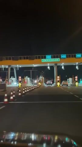A nighttime POV shot from inside a car moving toward a toll plaza. The sign above reads "İZMİR GİŞELERİ," which translates to "Izmir Toll Booths." The HGS system is indicated. Several lanes are visible, with traffic cones separating them. Traffic lights indicate which lanes are open. Other cars pass through the toll booths. Some lights reflect off the car's interior.