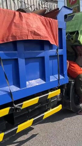 A person wearing a green shirt and dark pants is seen checking under the hood of a blue dump truck. The person is holding a plastic bottle of water in their right hand. The truck has a large blue cargo bed and yellow safety strips on the side. The background shows a corrugated metal wall. The scene appears to be outdoors during the daytime.