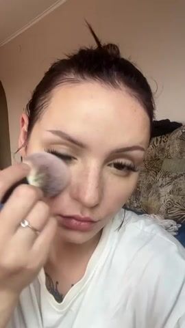 The media file is a close-up video of a young woman applying makeup. She has her hair pulled up in a bun, with loose strands framing her face. She is wearing a white t-shirt. She is holding a makeup brush in her right hand and applying powder to her cheek. Her expression is focused and serious as she concentrates on her makeup application. She has distinct freckles across her nose and cheeks, and wears long, false eyelashes. She is wearing a ring on her left hand. The background is softly blurred, showing what appears to be a bed with patterned pillows and a hint of a wall. The lighting suggests it is daytime, possibly indoors. The setting appears to be a bedroom in a home. No text is visible in the media file.