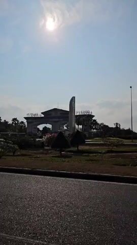 The video shows a road surrounded by dry grassy land and some trees and flowering bushes. The road is paved with black asphalt and marked with white lines. There is a median in the middle and a curb dividing the road from the dry grassy area. In the distance, there's what appears to be an elaborate gateway or entrance structure with flags. The sky is blue with scattered clouds, and the sun is shining brightly. As the video progresses, the view shifts down the road, revealing billboards and lamp posts lining the roadside.