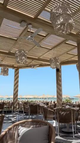 The video shows an outdoor restaurant setting. The roof is a wooden pergola with white macrame chandeliers and white ceiling fans. Below the pergola, there are round wicker chairs with grey cushions around tables on a patterned black-and-white tile floor. Beyond the seating area, there is a beach with straw umbrellas and a pier extending into the turquoise water under a clear blue sky.