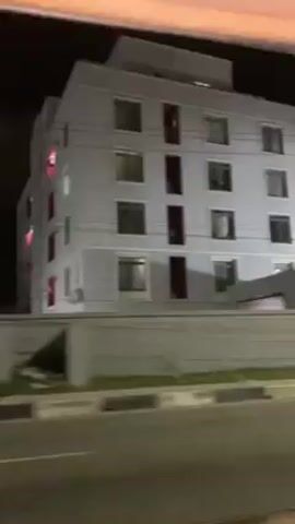 The video shows the view from a moving vehicle at night. The vehicle is traveling past a pair of four-story apartment buildings with gray walls and dark-framed windows, some with a red accent piece. There is a low wall running alongside the road. Streetlights illuminate the road. Beyond the buildings, a black wall is visible. The moving vehicle goes past a display of light projections, and an outdoor event venue, with flags and a digital billboard showing a colorful, abstract image. The scene is brightly lit.