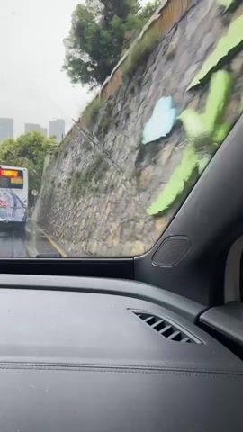 The video shows a view from inside a car, looking out through the windshield. It appears to be raining, as streaks of water are visible on the glass. A bus with "7801" displayed on its rear is visible in the distance, traveling on a road alongside a stone retaining wall. A circular speed limit sign with "20" is also visible on the side of the road. Trees and buildings can be seen beyond the bus. The interior of the car, including the dashboard with air vents and a speaker, is also visible in the foreground.