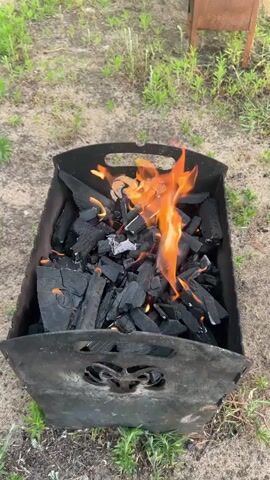 The video shows a close-up, overhead shot of a metal barbecue grill filled with charcoal. The charcoal is actively burning, with flames flickering and dancing amongst the black pieces. The grill itself appears to be made of dark, somewhat worn metal and has handles on the sides. The background consists of dry, sandy ground with patches of sparse green grass and some blurry foliage, suggesting an outdoor setting. There are no people or other objects clearly visible in the frame. The camera remains static throughout the short clip.