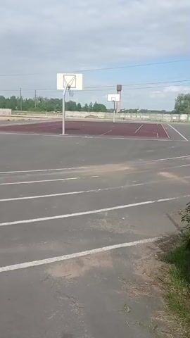 The video shows a view of an empty outdoor basketball court and running track. The basketball court has a dark red surface and two basketball hoops, both with white backboards and silver poles. The track area in the foreground is gray with white lines marking lanes. Beyond the court, the scenery includes a few trees and power lines against a partially cloudy sky. The camera appears to be handheld, creating a slightly unsteady perspective.