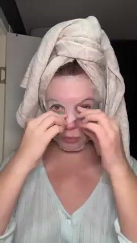 A woman in a light blue shirt and with a towel wrapped around her head is applying a facial sheet mask in front of a camera. She's pressing the mask around her nose, forehead, eyes, and cheeks, ensuring it adheres to her skin. A partially visible white cabinet is on the left and a dark space on the right.