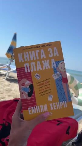 The video opens with a close-up shot of a book held in a person's hand. The book is titled "КНИГА ЗА ПЛАЖА" in large yellow letters against a red background, with the author's name "EMILY HENRY" printed below. The book cover features illustrations of people on a beach. The background is blurred but suggests a sunny beach scene. The camera then slowly pans away from the book, revealing the wider environment. The scene is a sandy beach on a clear, sunny day. The ocean is visible in the background with several people swimming and enjoying the water. Beach chairs and umbrellas are set up along the shore. In the foreground, a red towel with black writing is draped over a beach chair, and a green and white inflatable ring is also visible. The overall impression is a relaxing day at the beach with a book.