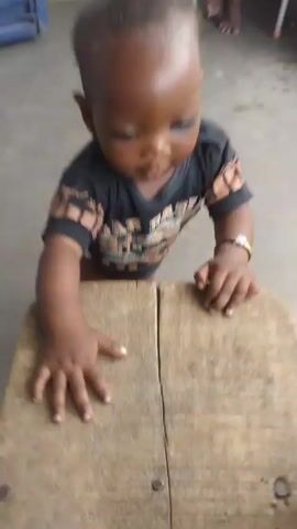 A baby with dark skin and short, curly hair is standing on a tile floor and holding onto a small, wooden stool. The baby is wearing a black T-shirt with gold accents and a gold bracelet on their left wrist. The stool is made of two planks of wood joined together. Behind the baby, a person is sitting in a chair and looking at a phone. There is a blue door visible in the background.