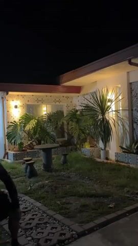 A short video shows an outdoor area at night. The scene is illuminated by artificial lights. In the foreground, there is a paved walkway and a grassy area with plants. To the right, there is a building with large windows and decorative tile accents. Lights are visible both inside and outside the building. Further into the background, there is a covered structure with a vehicle and more vegetation. The sky is dark, indicating it is nighttime.