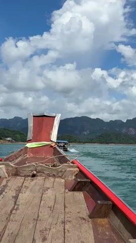 The video shows the perspective from the front of a wooden boat traveling on a body of water. The boat has a weathered wooden deck and a red and silver ornate structure at its front. In the distance, a second boat is visible. The background features lush green mountains under a bright blue sky with scattered white clouds. The water is choppy and appears to be a vibrant turquoise color.